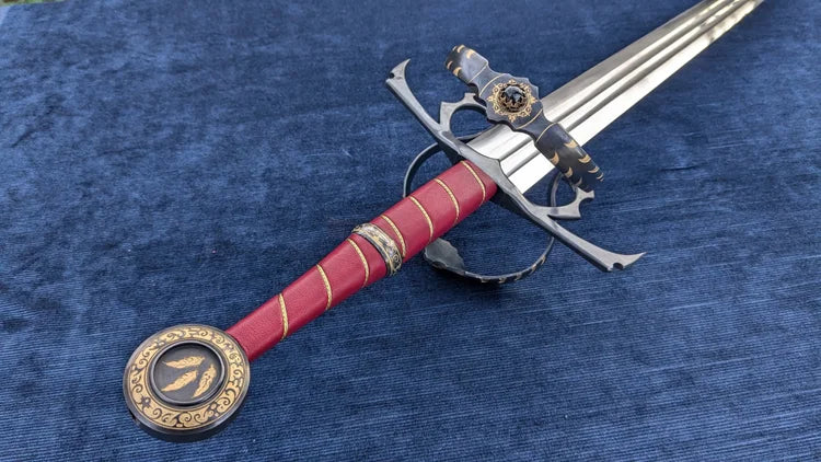 A decorative medieval-style sword with a red grip, gold accents, and a silver blade, displayed diagonally on a textured blue fabric background.