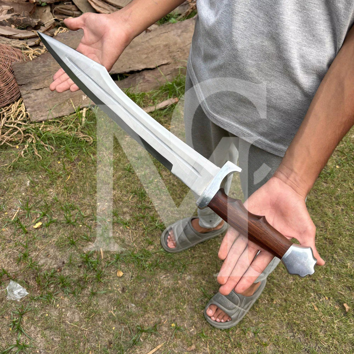 A person outdoors holding a large medieval-style sword with a silver blade and brown wooden handle, standing on grass and wood logs.