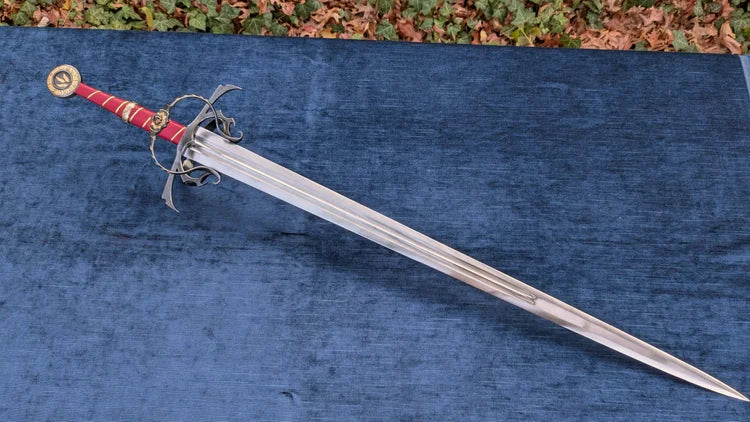 A medieval-style sword with a silver steel blade, a red wrapped hilt, and a black crossguard, displayed diagonally on a textured blue fabric surface.