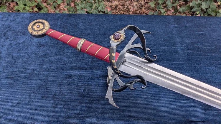 A decorative fantasy sword with a red grip, gold accents, and a silver blade, displayed diagonally on a blue textured fabric surface with outdoor foliage visible in the background.