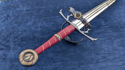 A decorative medieval-style sword with a red grip, gold accents, and a silver blade, displayed diagonally on a textured blue fabric background.