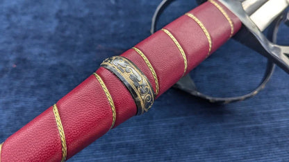 A close-up of a decorative sword hilt wrapped in textured red leather with gold braided stripes and an ornate black-and-gold metal guard, resting on a blue textured fabric background.