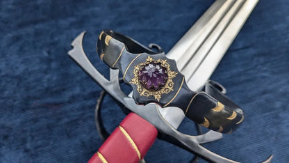 A decorative sword with a silver-colored blade, a black and gold hilt featuring a central purple gemstone, and a red wrapped grip, displayed on a textured blue fabric background.