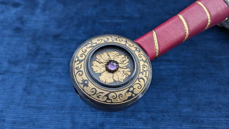A close-up of a decorative circular object with ornate gold scrollwork on a dark base, featuring a central floral motif and purple gemstone, resting on a textured blue surface next to a red handle with gold bands.