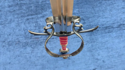 A close-up of a decorative sword hilt with a polished silver guard and pommel, a red wrapped grip, and a light-colored blade, set against a textured blue background.
