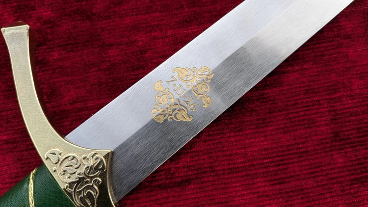 A close-up of a decorative dagger or sword blade with an engraved gold floral motif, resting on a textured red velvet surface. The hilt features ornate gold-toned metalwork and a dark green wrapped handle.