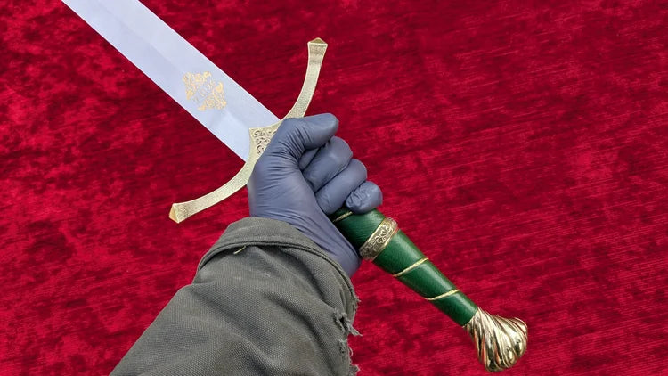 A hand wearing a blue nitrile glove holds a decorative sword with a green handle, gold guard and pommel, and a silver blade against a red velvet background.