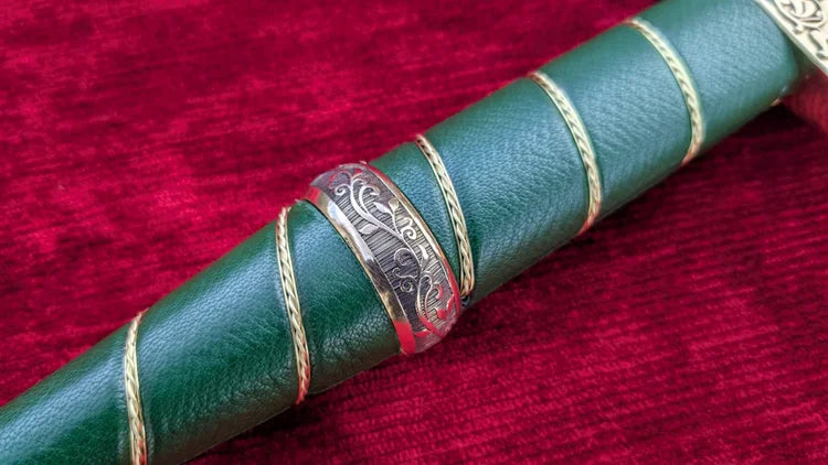 A close-up of a green leather-wrapped cylindrical product, likely a decorative handle or sleeve, featuring gold braided rope accents and a central silver-toned engraved band, set against a deep red velvet background.