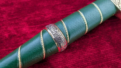 A close-up of a green leather-wrapped cylindrical product, likely a decorative handle or sleeve, featuring gold braided rope accents and a central silver-toned engraved band, set against a deep red velvet background.