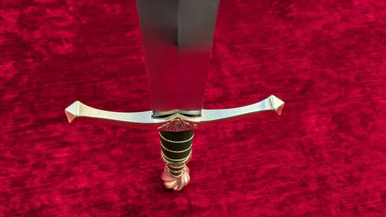 A close-up of a sword hilt and blade resting on a textured red velvet surface. The blade is silver, the crossguard and pommel are gold-toned, and the grip is wrapped in black cord with gold trim.