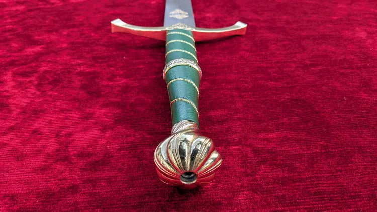 A decorative sword hilt and upper blade shown diagonally on a textured red velvet background. The hilt features a green wrapped grip with gold accents and a sculpted gold pommel.