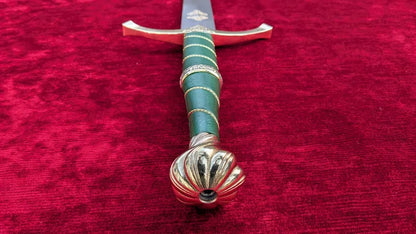 A decorative sword hilt and upper blade shown diagonally on a textured red velvet background. The hilt features a green wrapped grip with gold accents and a sculpted gold pommel.