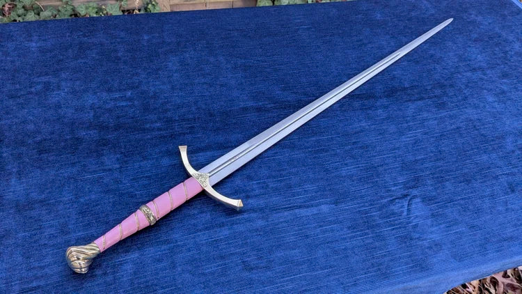 A medieval-style sword with a silver blade, gold guard and pommel, and a pink wrapped grip, displayed diagonally on a textured blue fabric surface.