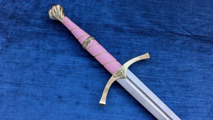A decorative sword with a pink wrapped handle, gold-colored metal fittings, and a silver blade, displayed diagonally on a textured blue fabric background.