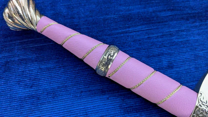 A close-up of a pink wrapped sword or dagger handle with gold braided cord accents and a silver engraved guard, resting on a textured blue background.