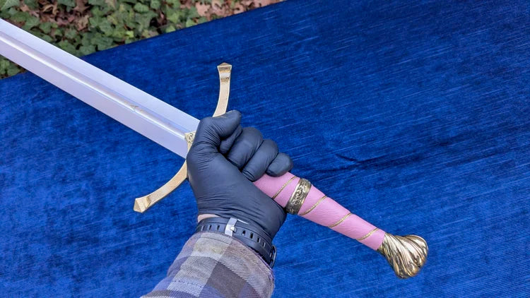 A close-up of a hand wearing a black glove holding a decorative sword with a silver blade, gold guard and pommel, and a pink wrapped grip, set against a bright blue textured background.
