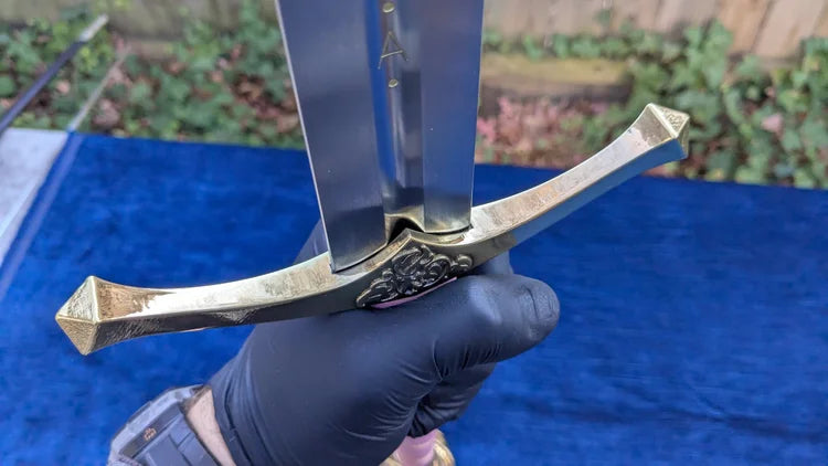 A close-up of a hand wearing a black glove holding a silver-colored sword with a decorative crossguard and a blue cloth background.