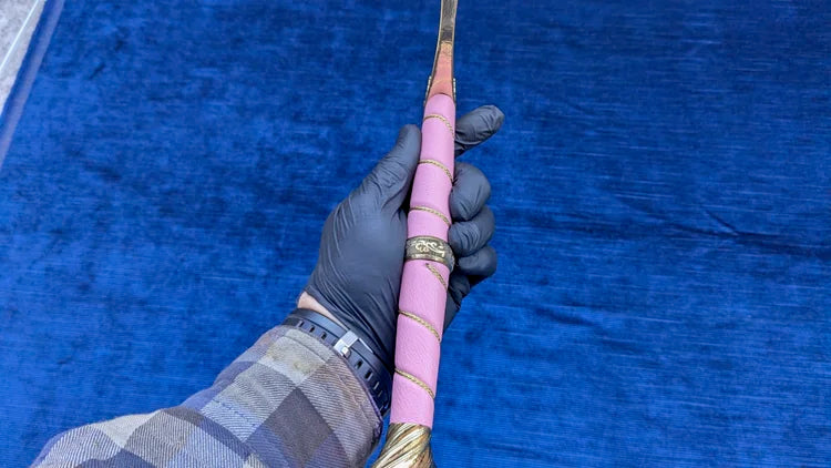 A close-up of a hand wearing a black glove holding a decorative sword or dagger with a pink wrapped handle and gold-colored metal fittings, set against a textured blue background.
