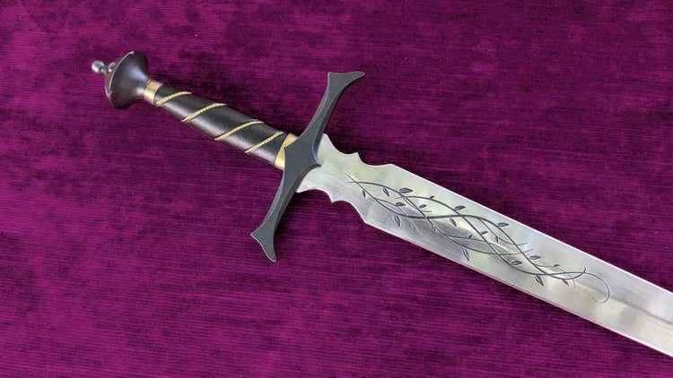 A decorative medieval-style sword with a silver engraved blade, black crossguard and pommel, and a black wrapped grip with gold spiral accents, displayed diagonally on a textured purple fabric background.