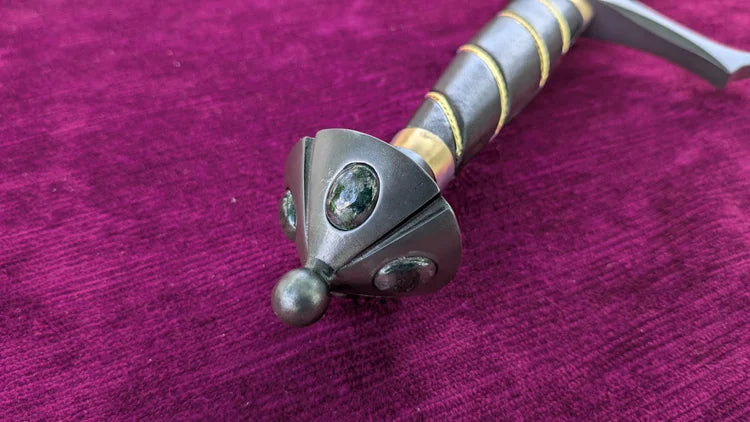 A close-up of a decorative dagger or sword hilt with a dark metal guard, oval green gemstones, and gold accents, resting on a textured magenta-purple velvet surface.