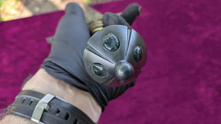 A close-up of a hand wearing a black glove holding a decorative metal object with four dark oval stones, set against a bright magenta-purple textured background.