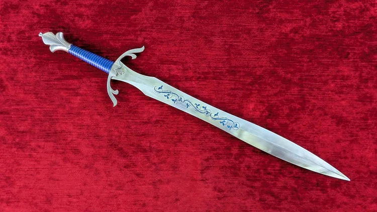 A decorative fantasy dagger with a silver blade featuring blue engravings, a blue wrapped handle, and a silver crossguard, displayed diagonally on a textured red velvet background.