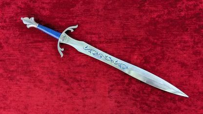 A decorative fantasy dagger with a silver blade featuring blue engravings, a blue wrapped handle, and a silver crossguard, displayed diagonally on a textured red velvet background.