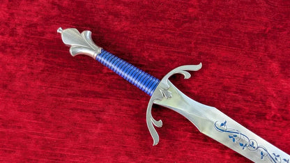 A decorative fantasy sword with a silver-colored blade and guard, a blue wrapped hilt, and blue floral engravings, displayed diagonally on a textured red velvet background.
