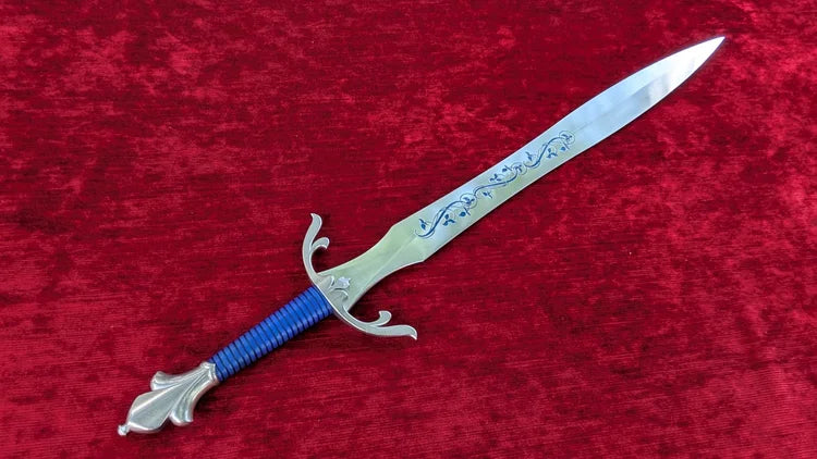 A decorative fantasy dagger with a silver engraved blade, a blue wrapped grip, and a silver hilt, displayed diagonally on a textured red velvet background.