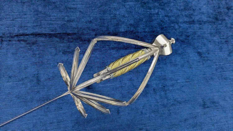 A metallic medical instrument, likely a catheter or stent-related device, featuring a silver metal frame, a yellow-gold coiled internal element, and a white plastic connector, shown on a textured blue background.