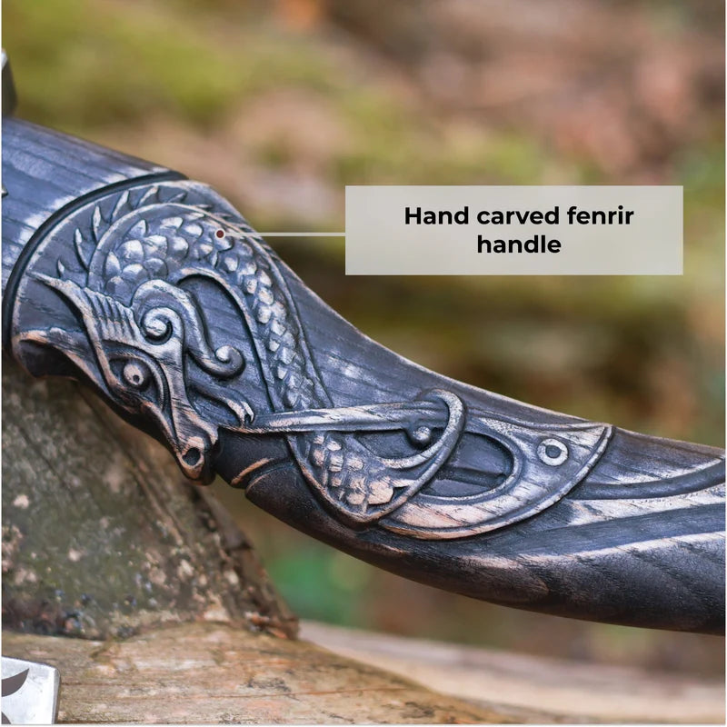 Close-up of a hand-carved dark metal or wood handle with an ornate dragon-like carving, shown outdoors on a log with a text label reading 'Hand carved fenrir handle.'.