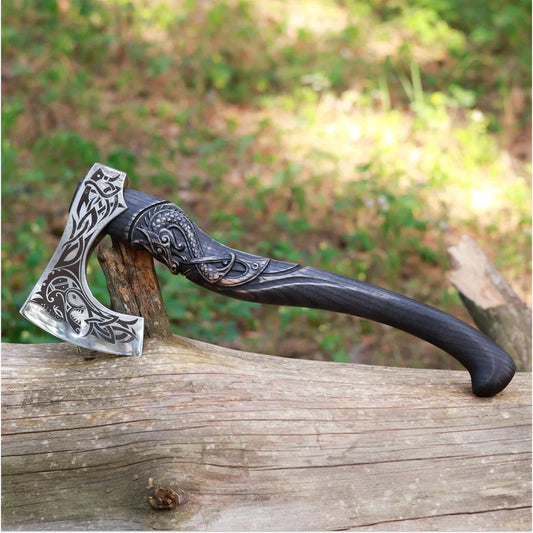 A decorative axe with an ornate engraved silver-toned head and a dark carved handle, resting on a weathered log outdoors with blurred greenery in the background.