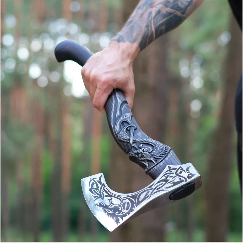 A close-up of a tattooed hand holding a decorative axe with an ornate dark handle and a silver blade featuring engraved Norse-style designs, set against a blurred forest background.