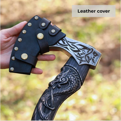A hand holds a decorative axe with an ornate dark carved handle and a black leather cover on the axe head. The background is a blurred outdoor scene, and a label in the top right reads 'Leather cover'.