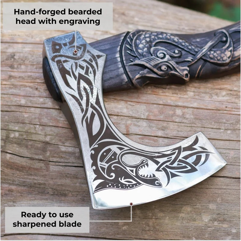A close-up of a hand-forged bearded axe head with engraved Norse-style animal and knotwork patterns, resting on a rustic wooden surface. Text overlays highlight the hand-forged head with engraving and the ready-to-use sharpened blade.
