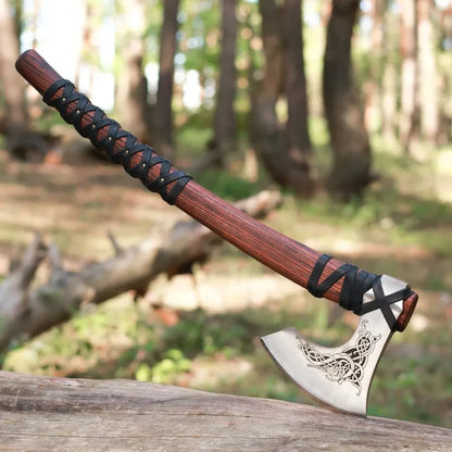 A hand axe with a dark brown wooden handle wrapped in black cord, resting on a log in a blurred forest setting.