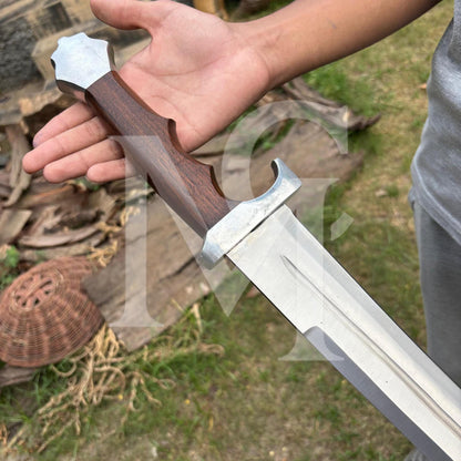 A close-up of a person's hand holding a medieval-style sword with a dark brown wooden handle and a silver steel blade, set against an outdoor rustic background.