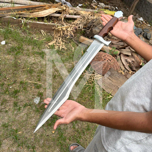 A person holding a large, custom-made training sword outdoors. The sword features a wide, silver-colored blade with a central groove, a brown wooden handle, and a silver guard.
