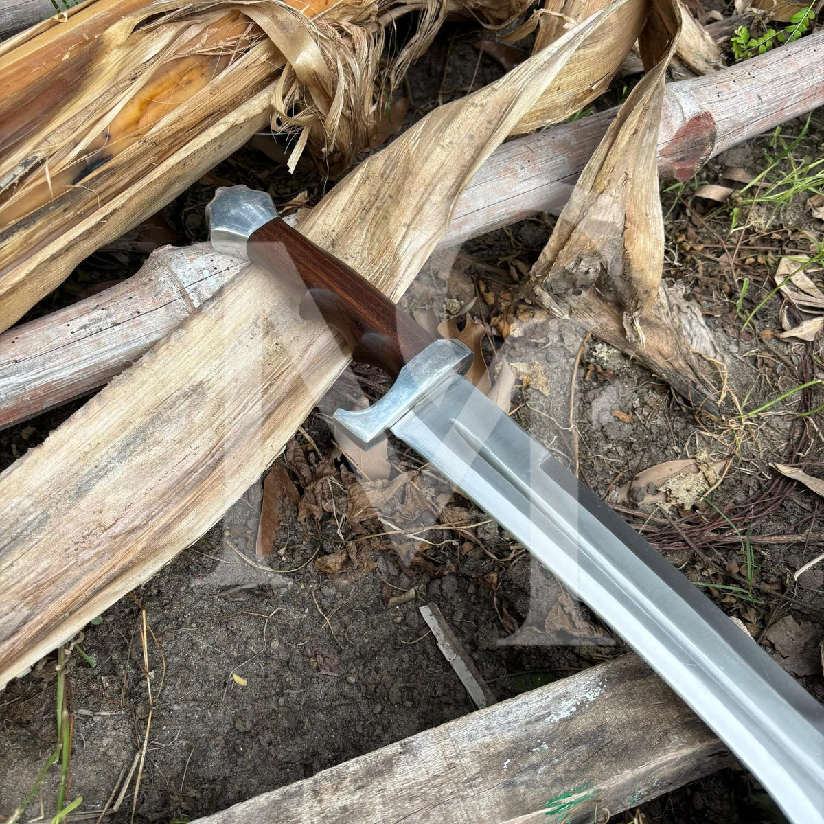 A medieval-style sword with a silver steel blade and a dark brown wooden hilt resting on a pile of dried corn husks and logs outdoors.