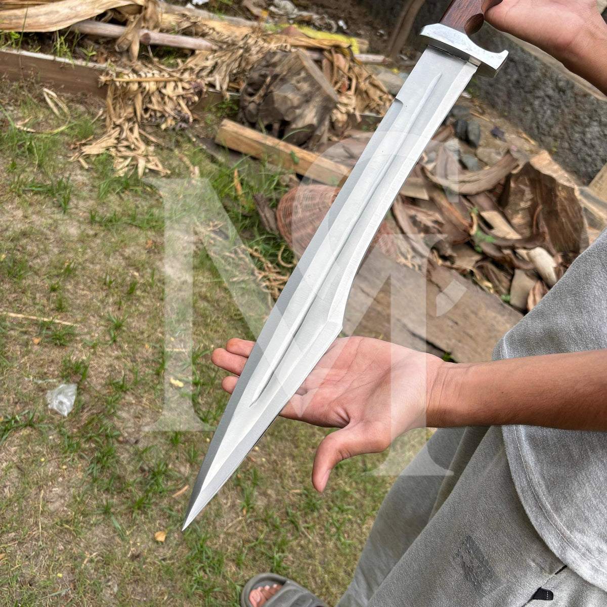 A person outdoors holding a large silver-colored machete or knife with a dark wooden handle. The background shows grass, dirt, and dried leaves.