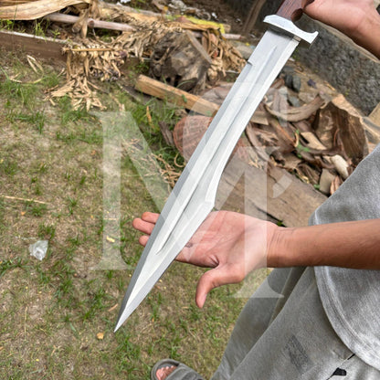 A person outdoors holding a large silver-colored machete or knife with a dark wooden handle. The background shows grass, dirt, and dried leaves.