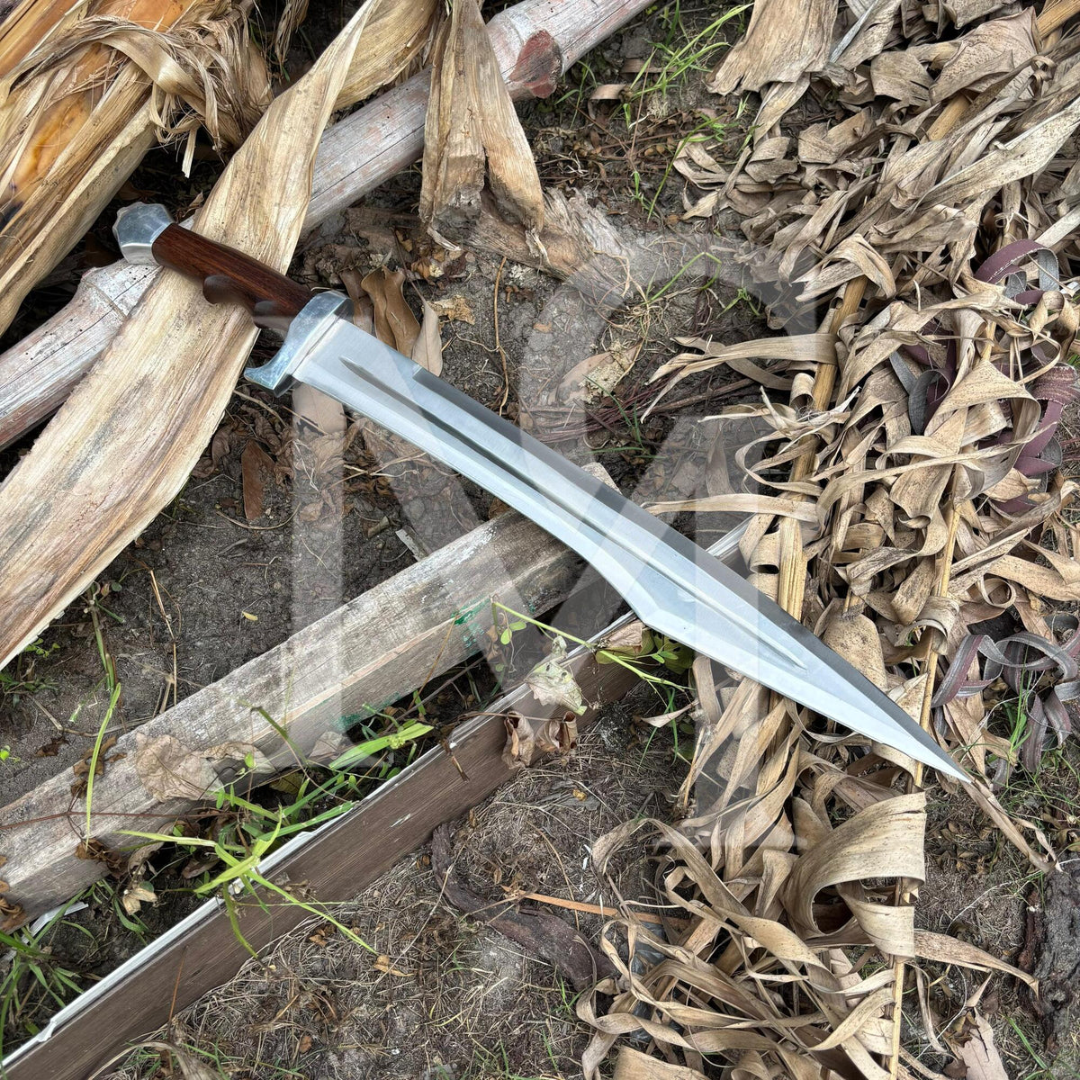 A fixed-blade knife with a silver steel blade and a dark brown wooden handle, resting diagonally on a pile of dried palm leaves and wood outdoors.