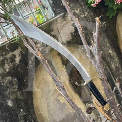 A curved sword with a silver blade, gold guard, and black wrapped handle, displayed diagonally among tree branches in front of a stone wall.