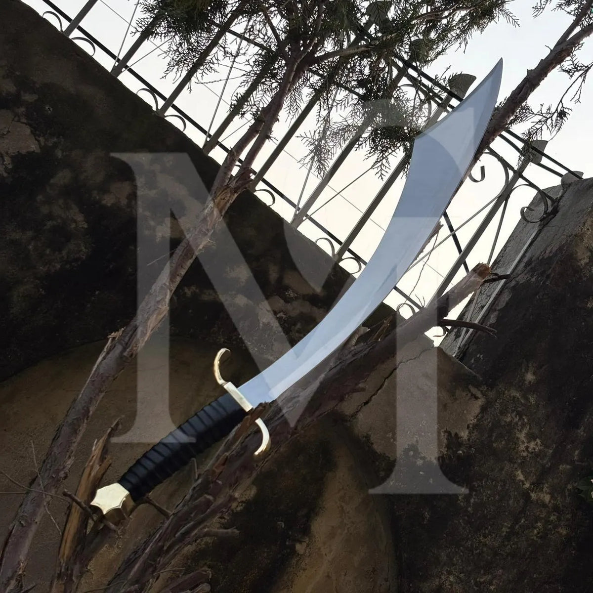 A curved sword with a silver blade, black wrapped handle, and gold guard, displayed outdoors against a rustic stone wall and metal fence.